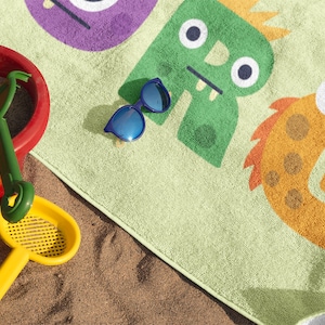 May include: A green and orange beach towel with a monster design. The towel is on a sandy beach with a red bucket and a yellow sand shovel.