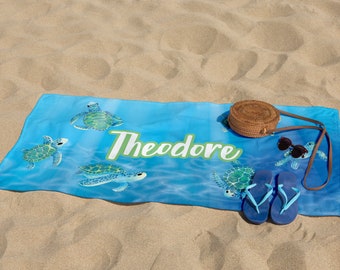 May include: A blue beach towel with a sea turtle design and the name "Theodore" printed in green. The towel is on a sandy beach with a straw bag and flip flops.