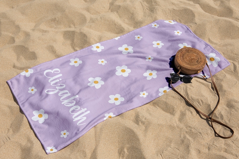 May include: A purple beach towel with a white daisy pattern and the name "Elizabeth" printed on it. The towel is lying on a sandy beach with a brown wicker bag and sunglasses.