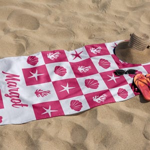 Puede incluir: Toalla de playa blanca con un patr&oacute;n de cuadros rosa y blanco. La toalla presenta dise&ntilde;os rosas de conchas marinas, estrellas de mar y corales. La palabra "Margot" est&aacute; impresa en negro en la toalla. Un sombrero de paja, gafas de sol y chanclas est&aacute;n sobre la toalla.
