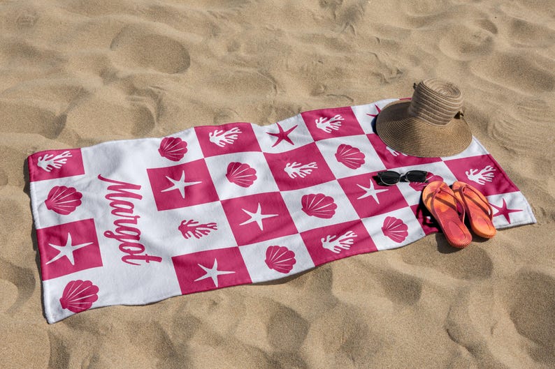 Puede incluir: Toalla de playa blanca con un patr&oacute;n de cuadros rosa y blanco. La toalla presenta dise&ntilde;os rosas de conchas marinas, estrellas de mar y corales. La palabra "Margot" est&aacute; impresa en negro en la toalla. Un sombrero de paja, gafas de sol y chanclas est&aacute;n sobre la toalla.