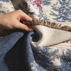 May include: A folded blue and white throw blanket with a detailed pattern. The blanket has a fringed edge and a design featuring trees and figures in red and blue. The blanket is made of a soft material.