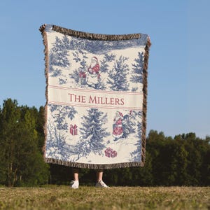 May include: A blue and white Christmas-themed throw blanket with a toile design featuring Santa Claus, gifts, and trees. The blanket has a fringed edge and the text "THE MILLERS" in red. The blanket is held up against a blue sky.