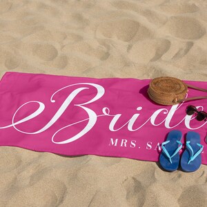 May include: Pink beach towel with the word "Bride" in white cursive lettering and the text "MRS. [last name]" below. The towel is on a sandy beach with a straw hat, sunglasses, and blue flip-flops.
