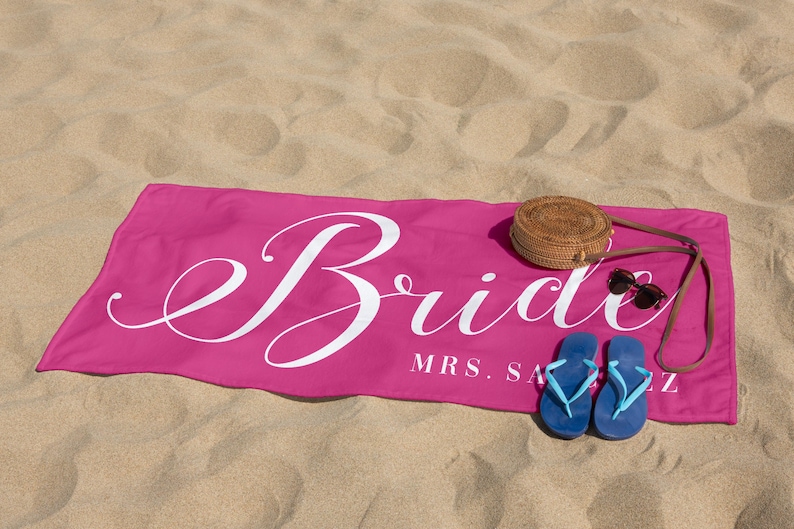 May include: Pink beach towel with the word "Bride" in white cursive lettering and the text "MRS. [last name]" below. The towel is on a sandy beach with a straw hat, sunglasses, and blue flip-flops.