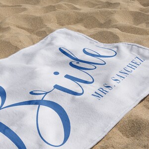 May include: White beach towel with the word "Bride" in blue cursive lettering and the text "Mrs. Sanchez" in smaller blue lettering.