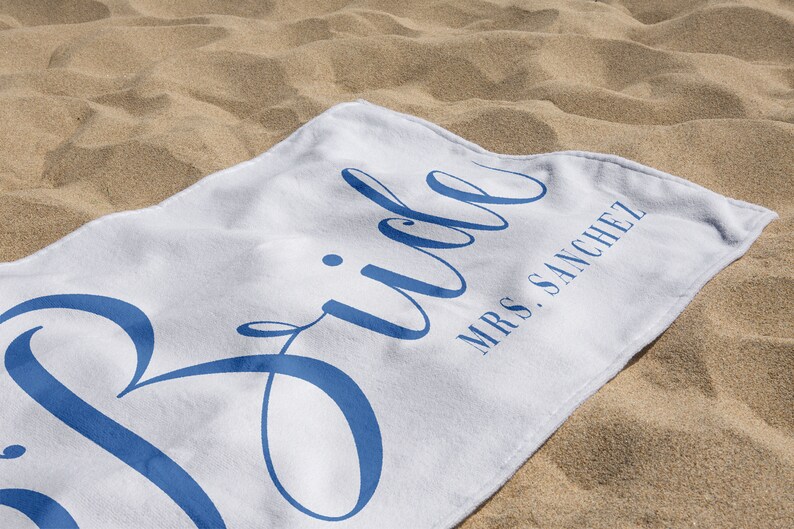May include: White beach towel with the word "Bride" in blue cursive lettering and the text "Mrs. Sanchez" in smaller blue lettering.