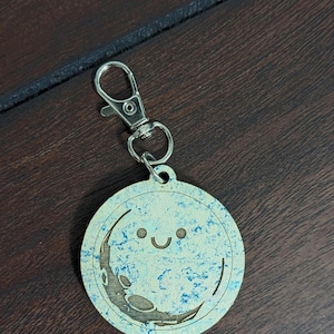 May include: A round keychain with a smiling moon design. The moon has a crescent shape and is light blue with darker blue accents. The keychain has a silver clasp and the letters "SRCL" are at the bottom.