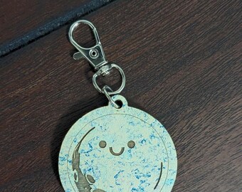 May include: A round keychain with a smiling moon design. The moon has a crescent shape and is light blue with darker blue accents. The keychain has a silver clasp and the letters "SRCL" are at the bottom.