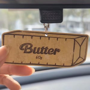 May include: A wooden rear view mirror accessory shaped like a butter stick. The word "Butter" is written in a stylized font, with "613g" below it. The accessory is hanging from a silver chain.