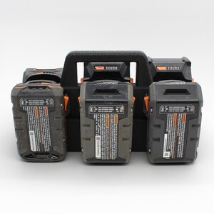 May include: A black battery organizer with a handle holds four black and gray power tool batteries. The batteries have orange accents and warning labels. The organizer is designed to hold the batteries securely.