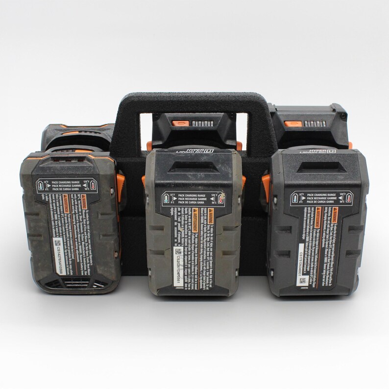 May include: A black battery organizer with a handle holds four black and gray power tool batteries. The batteries have orange accents and warning labels. The organizer is designed to hold the batteries securely.