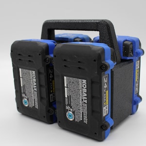 May include: A black and blue battery charger with a carrying handle. The charger has two 24V MAX 2.0 Ah KOBALT batteries attached to it. The batteries have a warning label that reads "WARNING/AVERTISSEMENT/ADVERTENCIA".