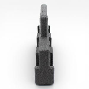 May include: A dark gray, textured, geometric object with a rectangular base and a series of stacked, rectangular blocks. The object has a rough, granular surface and is set against a white background.