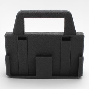 May include: A black, textured plastic object with a handle and rectangular cut-out. The object has a boxy shape with vertical and horizontal supports, and a textured surface. The design suggests a carrying case or holder.
