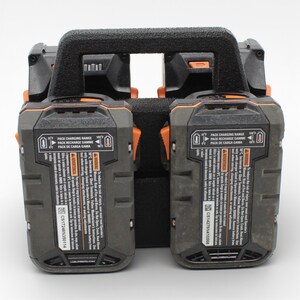 May include: A black and gray dual battery charger with an orange accent. The charger has a black handle and includes text that reads "PACK CHARGING RANGE" and "WARNING". The charger is designed for power tools.