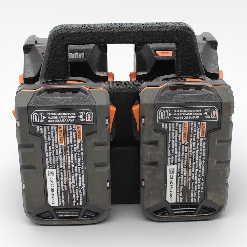 May include: A black and gray dual battery charger with an orange accent. The charger has a black handle and includes text that reads "PACK CHARGING RANGE" and "WARNING". The charger is designed for power tools.