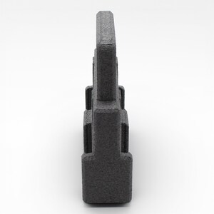May include: A dark gray, textured, rectangular object with a handle-like top. The object has a series of rectangular protrusions along its sides. The item is set against a white background.