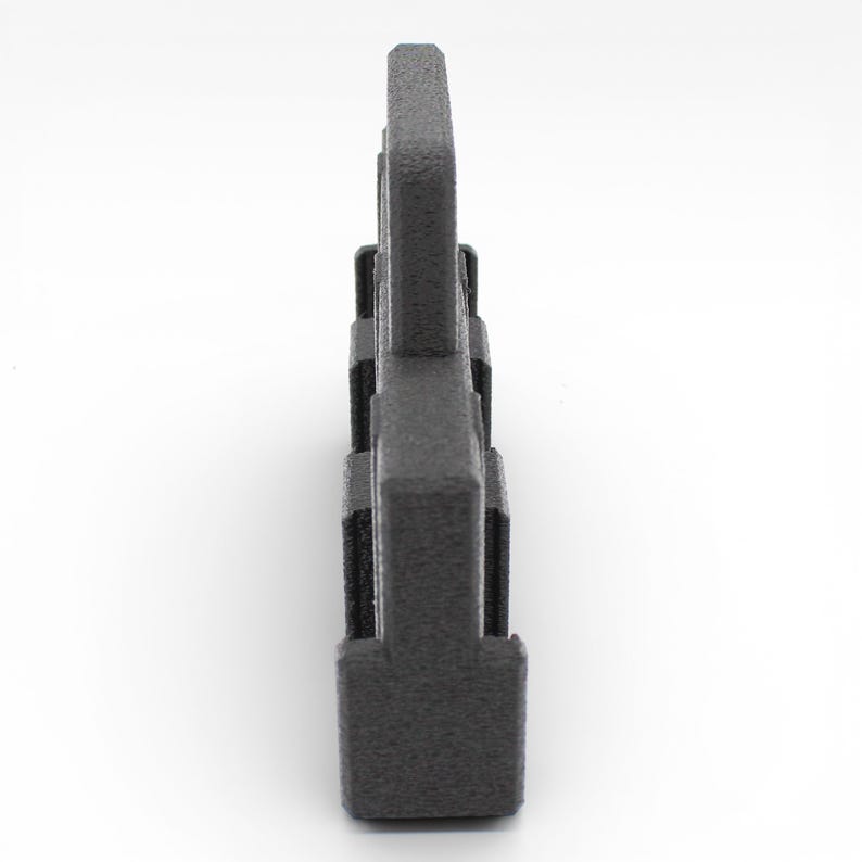 May include: A dark gray, textured, plastic object with a rectangular base and a series of vertical, rectangular protrusions. The object is viewed from the side against a white background. The overall shape resembles a stylized letter.
