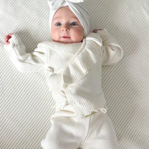 May include: A white knitted baby outfit with a ruffled top and matching pants. The baby is wearing a white bow headband.