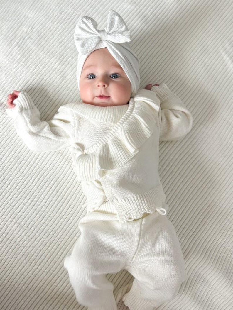 May include: A white knitted baby outfit with a ruffled top and matching pants. The baby is wearing a white bow headband.