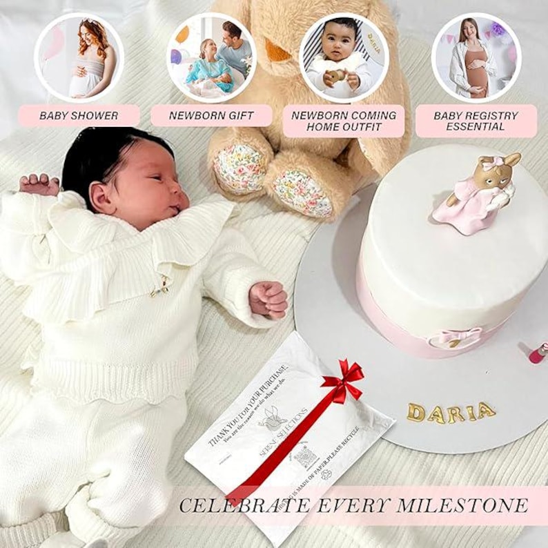 May include: A white baby outfit with a ruffled collar and a bow. The outfit is laid out on a white blanket with a white and red gift bag and a white cake with pink frosting and a small white figurine of a bear wearing a pink dress. The text "DARIA" is written on the cake.