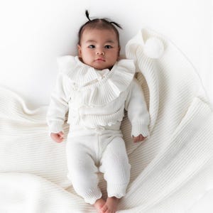 May include: A baby wearing a white knitted outfit with a ruffled collar and matching pants. The baby is lying on a white knitted blanket.