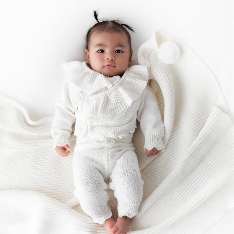 May include: A baby wearing a white knitted outfit with a ruffled collar and matching pants. The baby is lying on a white knitted blanket.