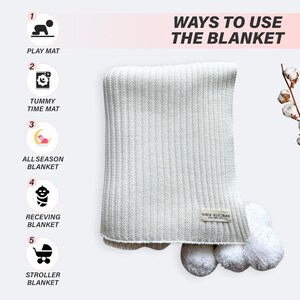 May include: A white knitted baby blanket with pom-poms on the corners. The blanket is folded and displayed on a white background. The text "WAYS TO USE THE BLANKET" is displayed above the blanket. Five icons are displayed on the left side of the image, showing different ways to use the blanket, such as a play mat, tummy time mat, all-season blanket, receiving blanket, and stroller blanket.