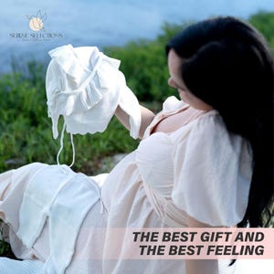 May include: A pregnant person holding a white knitted baby hat with ruffles. The text "The Best Gift and The Best Feeling" is on the bottom of the image.