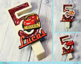 May include: A white number 5 candle with red glitter and a Lightning McQueen design. The name "Lucas" is written in red script. The candle is for a birthday celebration.