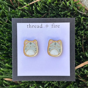 Pode incluir: Um par de brincos de madeira com um design de gato de desenho animado bonito. O gato é cinza com patas brancas e um nariz rosa. Os brincos estão sobre um fundo branco com o texto "thread + fire" sobre um fundo preto.