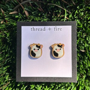 Peut inclure: Une paire de boucles d'oreilles en bois avec un motif de vache noir et blanc. Les boucles d'oreilles sont sur une carte blanche avec le texte "thread + fire" imprimé dessus.