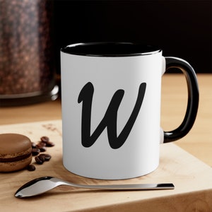 May include: White ceramic mug with a black interior and handle. The letter "W" is displayed in black cursive on the front. The mug sits on a wooden surface with a spoon, coffee beans, and a macaron.