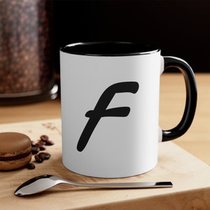 Può includere: Tazza in ceramica bianca con bordo e manico neri. La tazza ha una lettera "F" nera scritta a mano sul davanti.