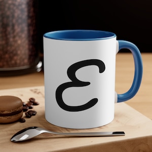 May include: A white ceramic mug with a blue rim and handle. The mug has a black stylized letter "E" printed on the front.