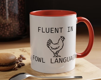 May include: White ceramic mug with a red rim and handle. The mug has black text that reads "Fluent in Fowl Language" and a black line drawing of a chicken.
