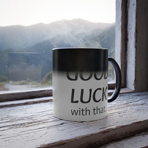 Op de afbeelding: Een zwart-witte kleurveranderende mok met de tekst "good luck with that." erop gedrukt. De mok is gevuld met een warme drank en er stijgt stoom uit de mok.