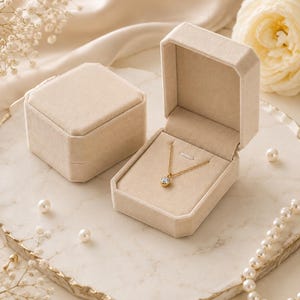 May include: A gold chain necklace with a single round clear stone pendant, presented inside an open beige velvet jewelry box. A second closed beige velvet jewelry box sits beside it, both featuring a soft, matte, light-colored fabric finish.