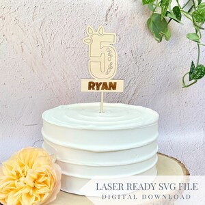 Könnte beinhalten: Ein Holz-Kuchentopper mit der Zahl 5 und dem Namen "RYAN" auf einem weißen Kuchen. Die Zahl 5 ist mit Blättern und Punkten verziert.