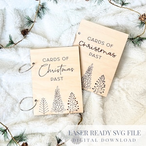 May include: Two wooden albums with silver ring binders, each with the words "CARDS OF Christmas PAST" and a decorative tree design. The albums are light-colored and rest on a white, fluffy surface with pine branches.