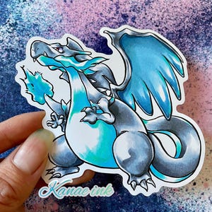 May include: A blue and gray cartoon sticker of a dragon with wings, a tail, and a blue flame coming out of its mouth. The sticker has the text "Kanae Ink" at the bottom.