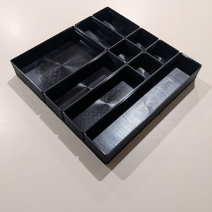 May include: A black plastic drawer organizer tray featuring multiple rectangular and square compartments of varying sizes. The tray has a structured grid design with smooth interior surfaces and vertical walls, providing organized sections for storage.