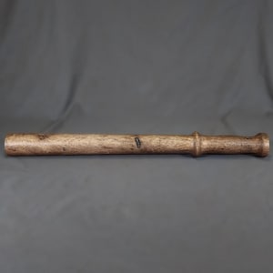 May include: A cylindrical wooden rolling pin with a dark brown finish and visible natural wood grain. The tool features a smooth, elongated body that tapers slightly towards one end, ending in a rounded handle section with a distinct raised ring.