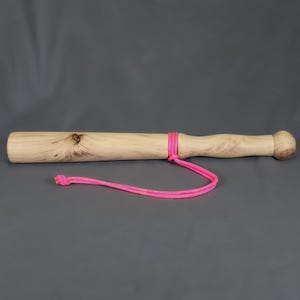 May include: A cylindrical wooden club with a smooth, light-coloured grain featuring natural knots. The handle section is contoured and ends in a rounded knob. A bright neon pink cord is tied around the centre, forming a loop with a knotted end.