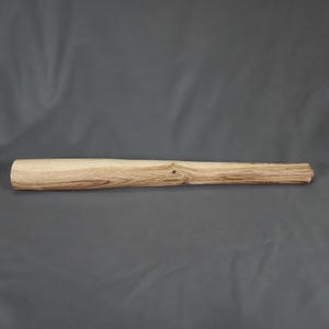 May include: A cylindrical wooden handle with a natural light brown grain pattern and a smooth, polished finish. The object features a slightly tapered shape, showcasing visible wood knots and organic textures along its length.