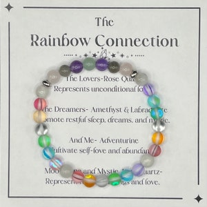 May include: A rainbow colored beaded bracelet with a white card that reads "The Rainbow Connection" and describes the meaning of each color bead. The Lovers-Rose Quartz represents unconditional love. The Dreamers-Amethyst & Labradorite promote restful sleep, dreams, and magic. And Me- Aventurine cultivate self-love and abundance. Moonstone and Mystic Quartz represent healing and love.