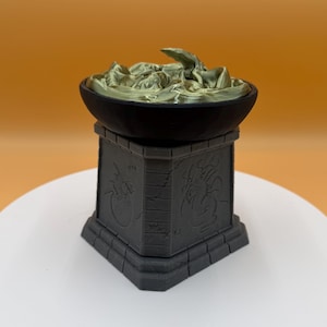 May include: A dark gray decorative pedestal with carved dragon designs supports a black bowl filled with a gold-colored, textured substance. The pedestal has a square base and a detailed, textured surface.