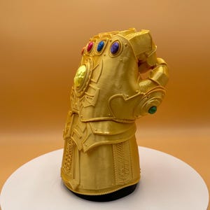 May include: A gold-colored gauntlet with six colorful gems. The gauntlet is detailed with intricate designs and is displayed on a white platform. The gems are red, blue, purple, yellow, and green.