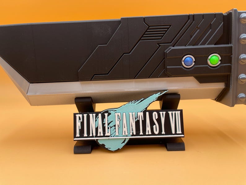May include: A black and silver Buster Sword replica from Final Fantasy VII, resting on a black stand. The sword features a detailed design with blue and green button accents. The Final Fantasy VII logo is displayed below.